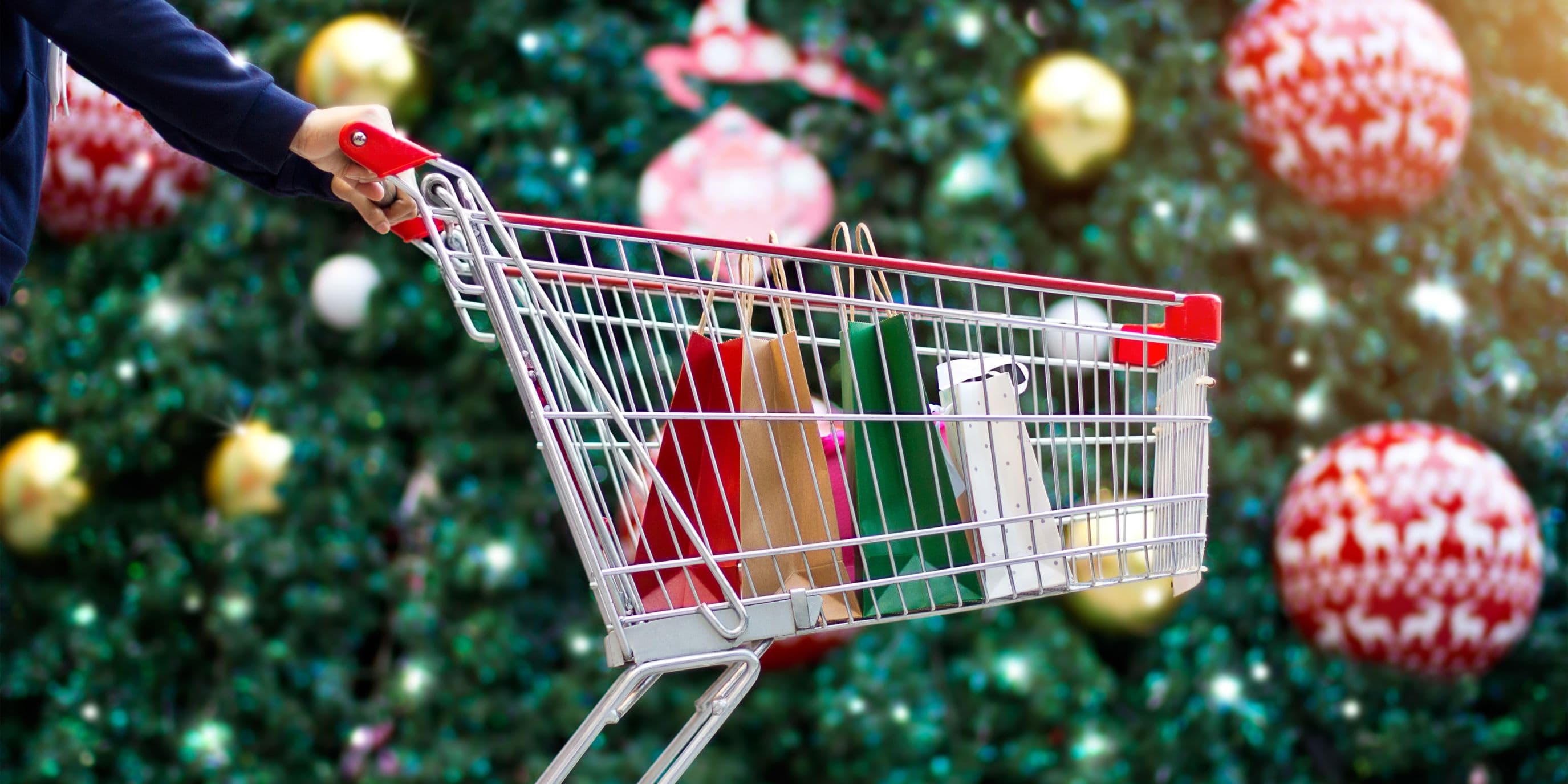 Mani che spingono un carrello pieno di regali di Natale. Sullo sfondo, un grande albero di Natale addobbato con palline di Natale